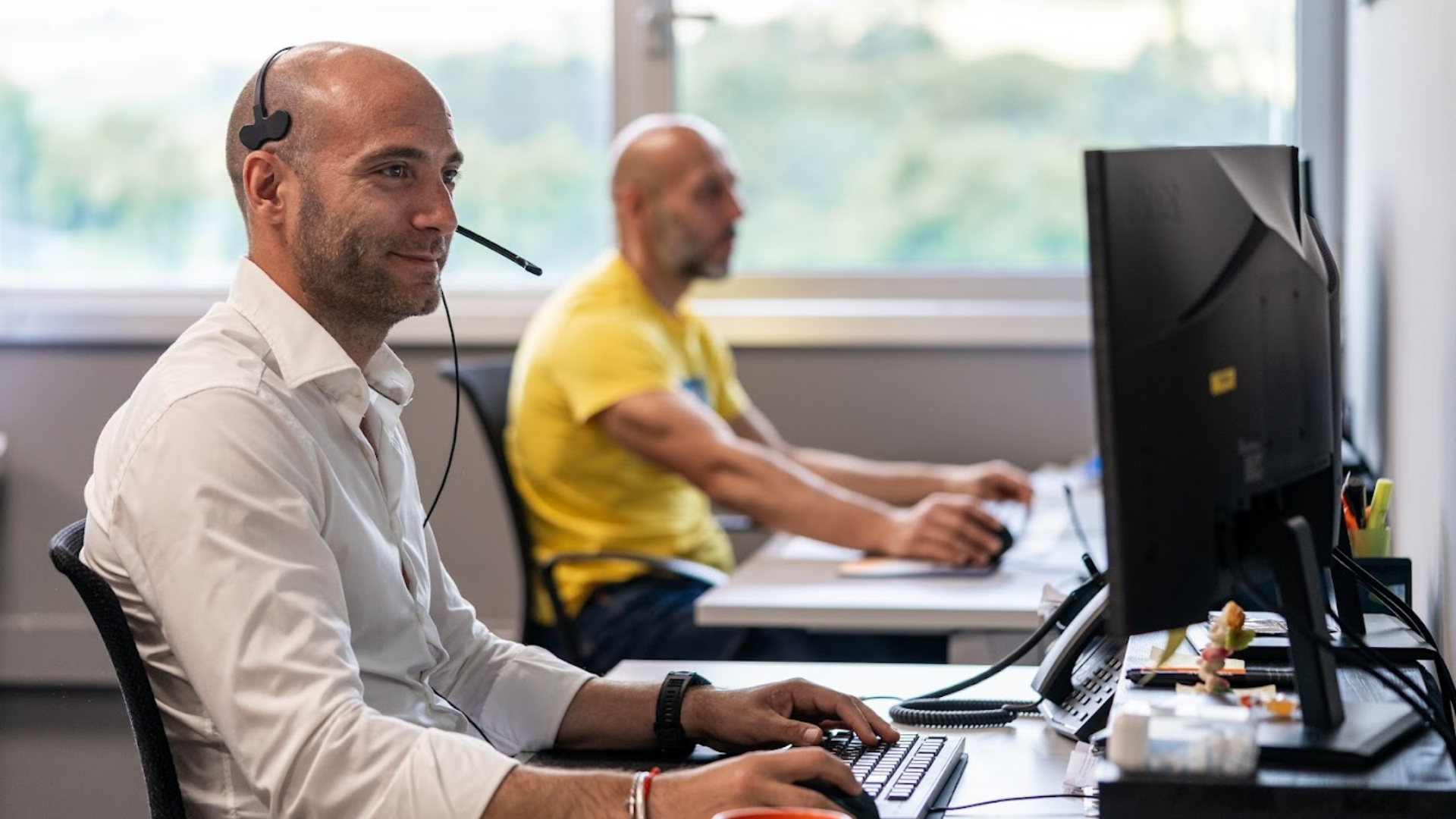 Due uomini lavorano al computer in un ufficio, uno indossa una cuffia con microfono.