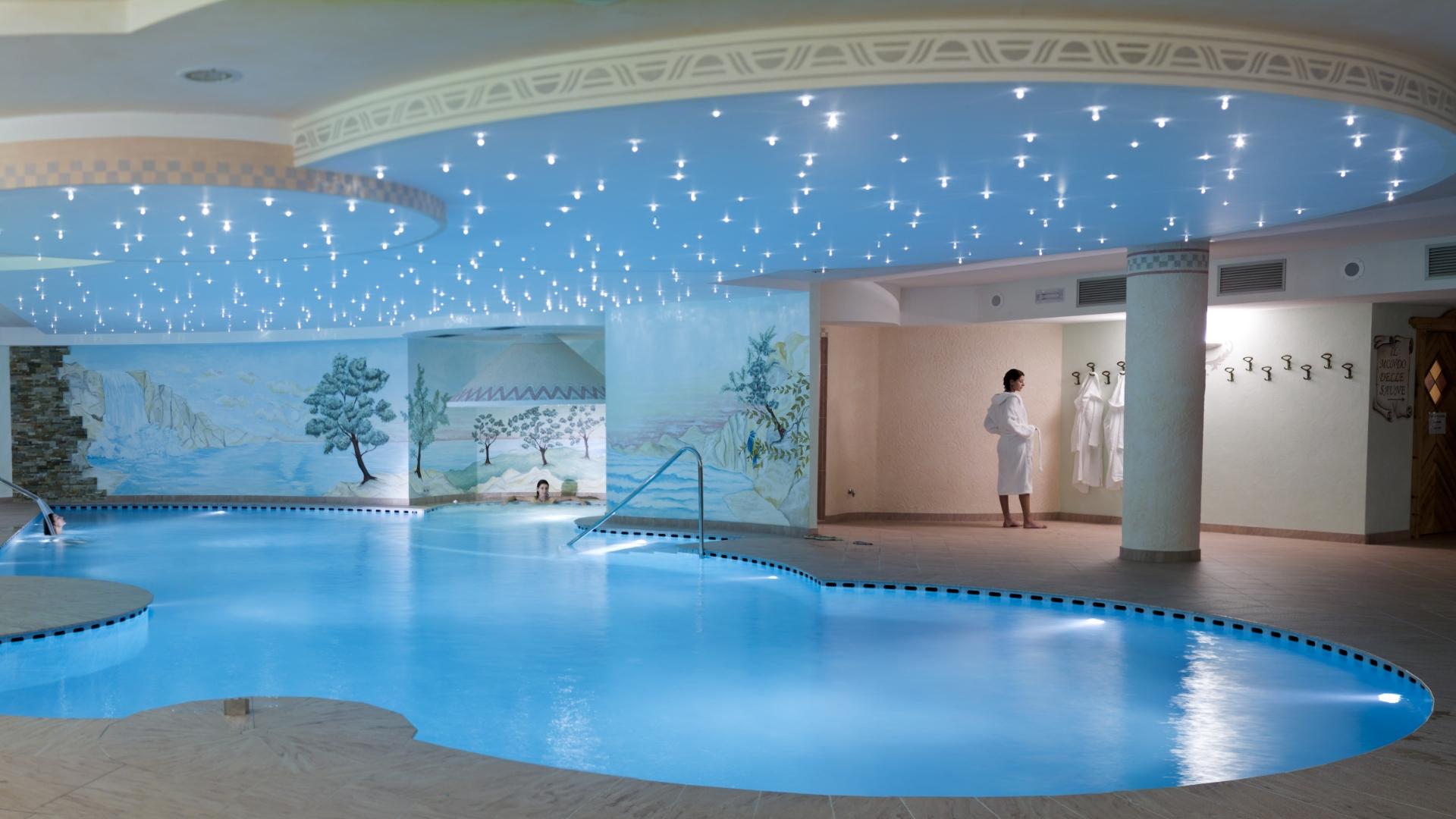 Piscina coperta con soffitto stellato e murales, persone in accappatoio.