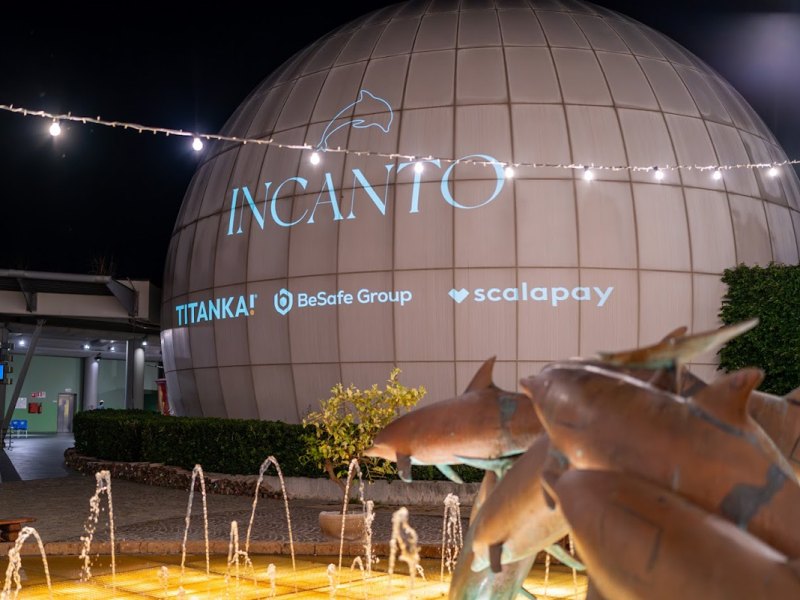 Esterno notturno di un edificio sferico con logo 'Incanto' e fontana con statue di delfini.