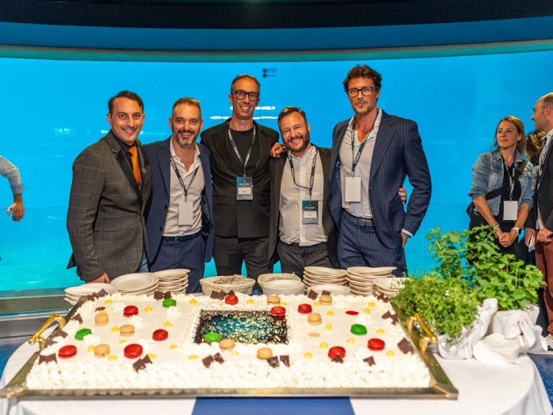 Gruppo di uomini in abiti formali davanti a una torta durante un evento.