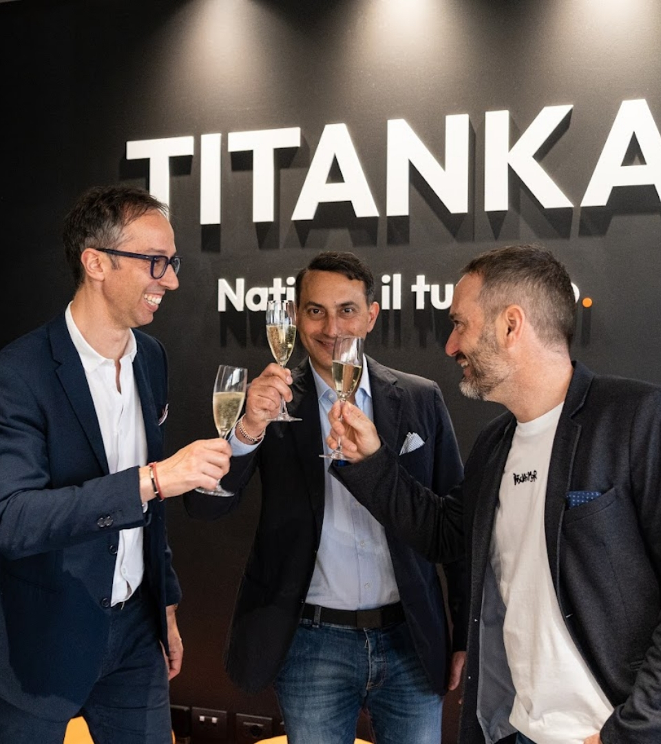 Tre uomini brindano davanti al logo TITANKA, sorridendo e tenendo bicchieri di champagne.